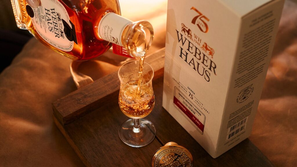 Close de cachaça Weber Haus âmbar sendo servida em copo de degustação, com embalagem ao fundo e luz dourada de fim de tarde.